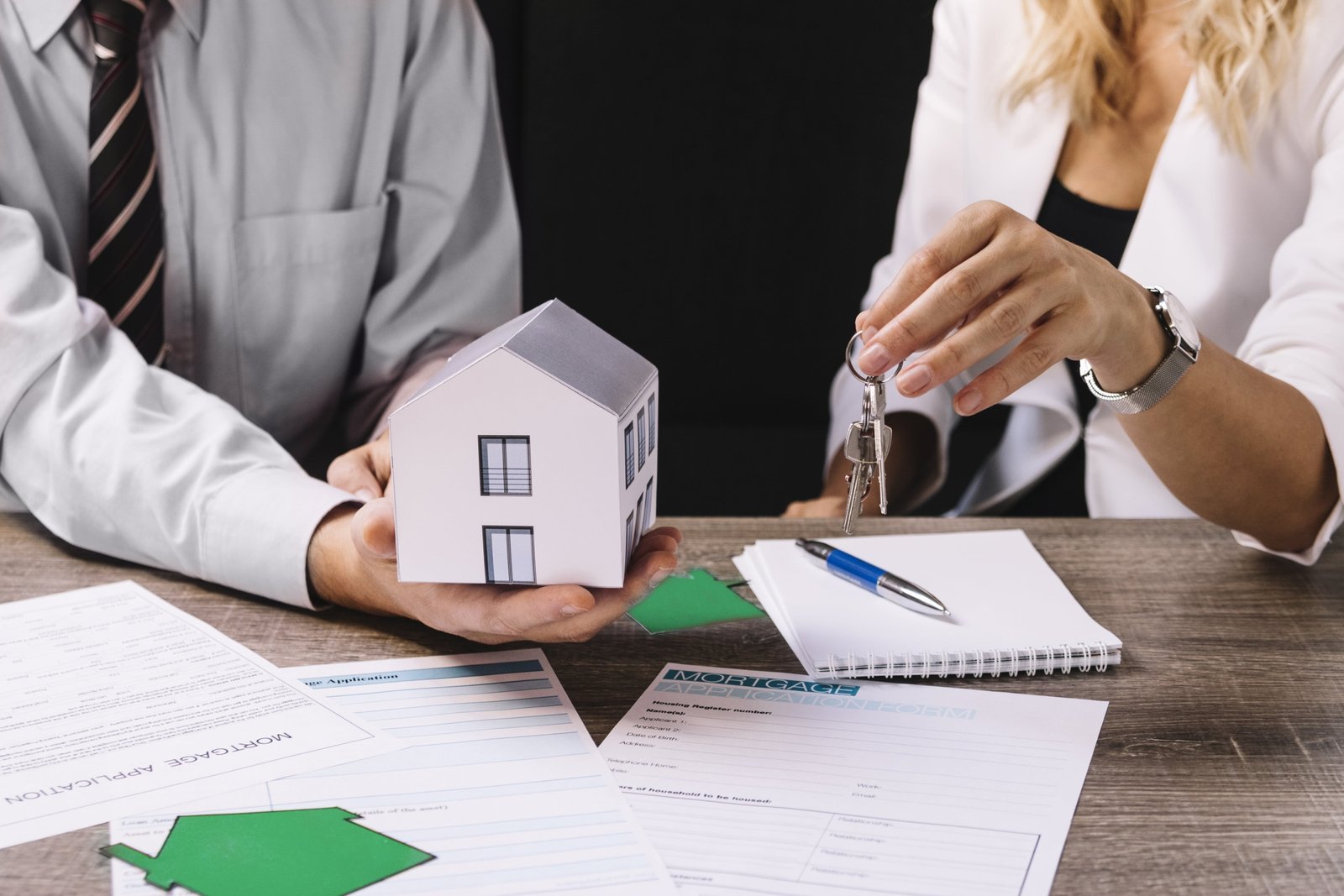 Two professionals sit at a desk reviewing real estate documents; one holds a small model house while the other gestures with glasses, suggesting a home buying or mortgage discussion.