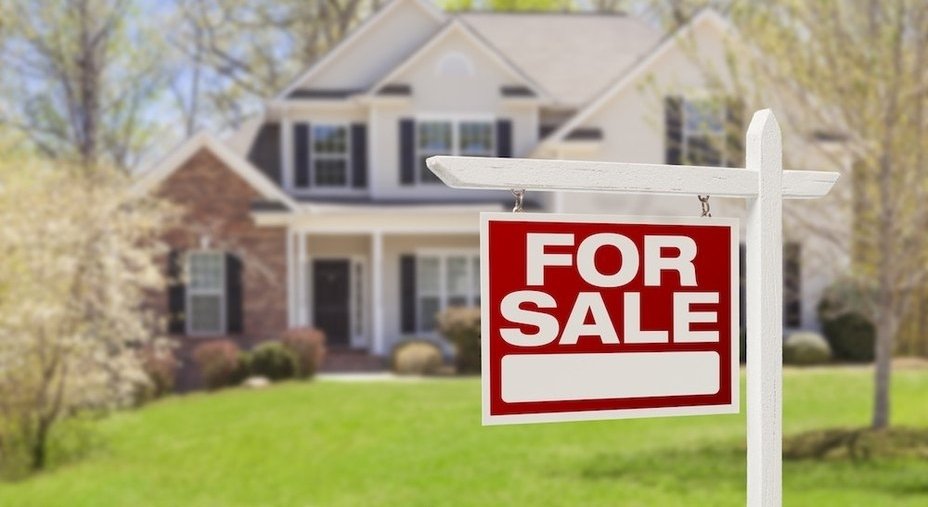 A “For Sale” sign stands in front of a large suburban house with a well-kept lawn and trees, representing a home listed on the real estate market.