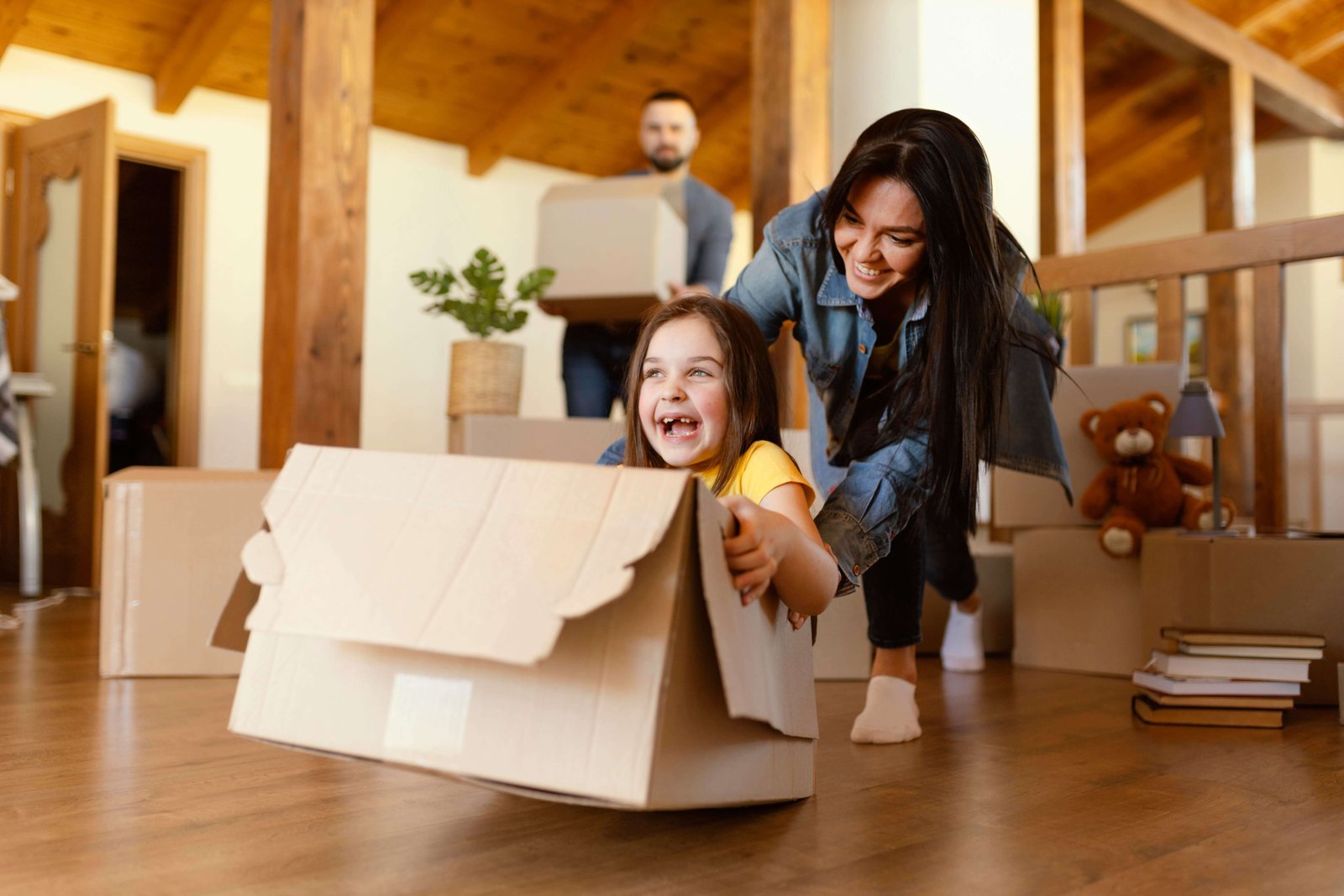 Happy family moving into a new home — a child laughing while sitting in a cardboard box being pushed by their mother, surrounded by moving boxes and warm wooden interiors.