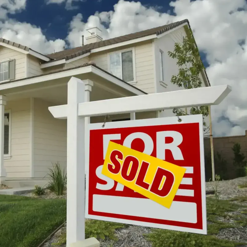 A “For Sale” sign with a yellow “Sold” sticker in front of a suburban house under a partly cloudy sky.